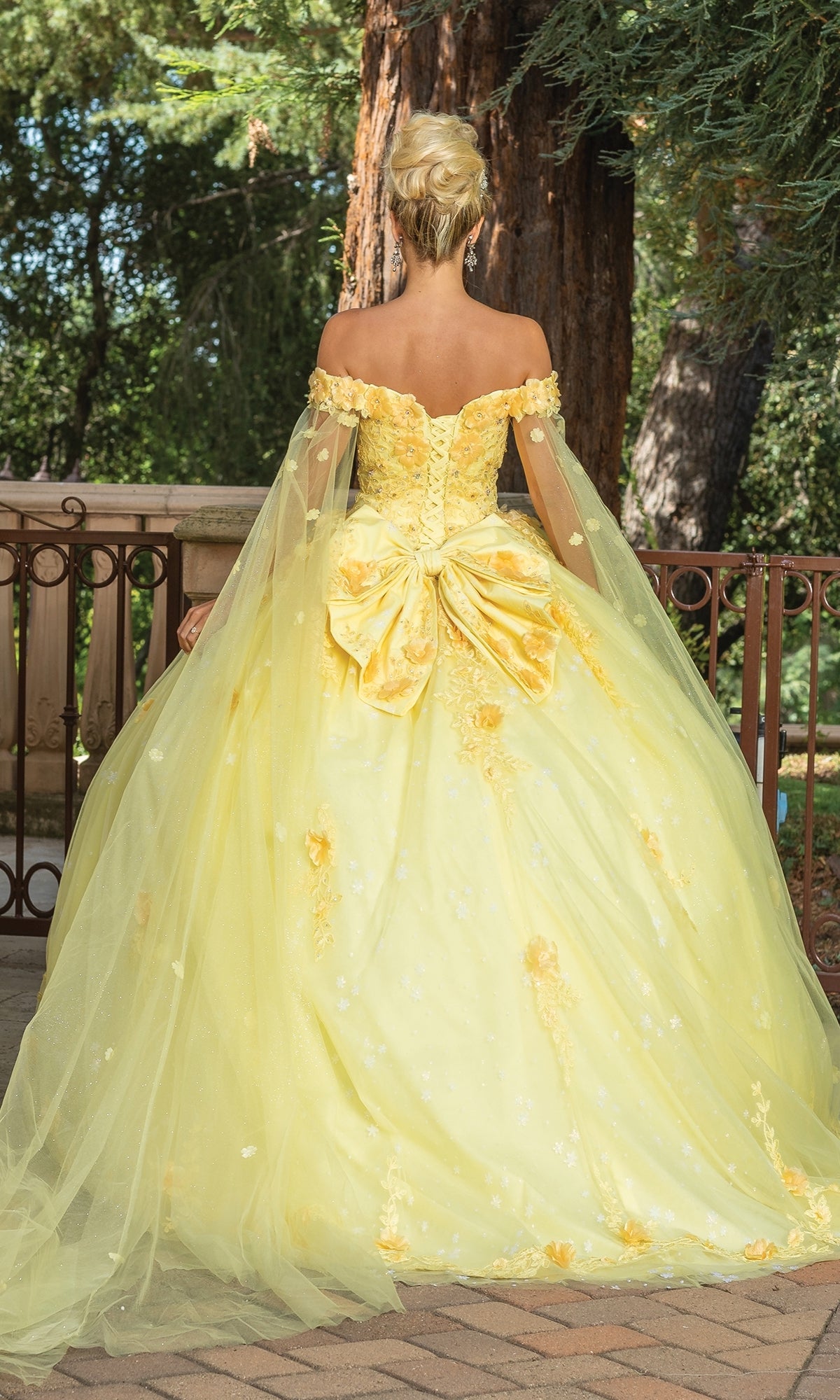 A woman stands outdoors amid lush greenery, wearing the Quinceanera Dress 1789 By Dancing Queen—an off-the-shoulder yellow ball gown featuring a large bow and tulle cape.